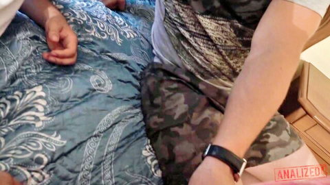 Guy in camo shorts sits on teal bed with hands reaching during threesome foreplay