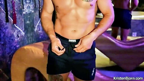 Muscular tattooed hunk gripping his tight black shorts waistband with ripped buddy in background at gay lounge