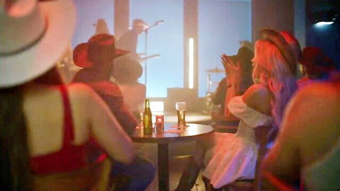 Sexy cowgirls in revealing tops and hats partying at neon-lit cowboy bar with beers and band.