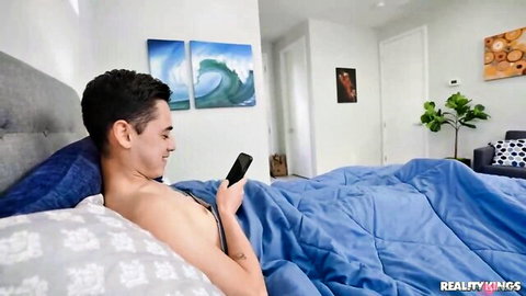 Young Latino man smiling in bed, topless under blue blanket, checking phone - stepmom creampie tease