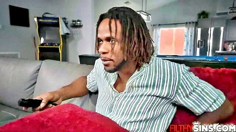 Dreadlocked black stud in striped shirt grips game controller on red couch intensely