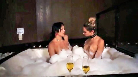 Two nude women in bubbly bath with champagne, one in collar for kinky fun