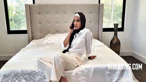 Seductive Arab woman in white hijab sits on bed, talking on phone