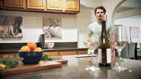 Curly-haired man in green shirt pouring white wine into glasses on kitchen counter