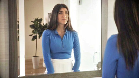 Aubry Babcock in blue zip-up top and white skirt, thoughtful mirror gaze
