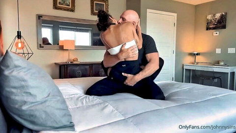 Muscular bald man passionately embraces brunette in white bra on his lap kissing intensely