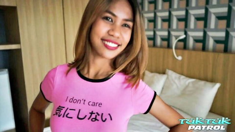 Cute tanned Asian babe smiling in tight pink 'I don't care' shirt teasing curves in bedroom