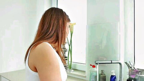 Brunette woman in tight white tank top leaning over bathroom sink