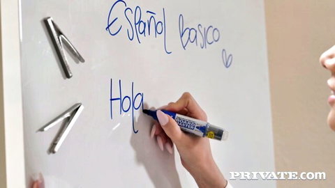Skinny Latina teacher with pink nails writing Hola on Español básico whiteboard