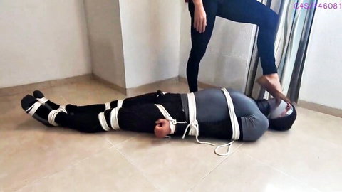 Femdom mistress stands over bound gagged male slave pressing bare foot on his roped body