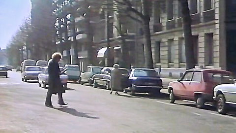 Curly blonde woman in long coat walks past parked 70s cars on vintage French street