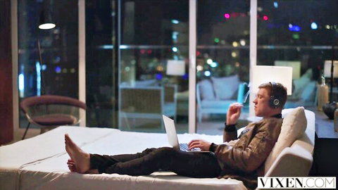 Handsome man in leather jacket lounges barefoot on bed typing on laptop with city night view