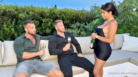 Sheila Ortega in short black dress with two men, champagne outdoors