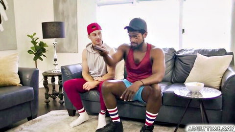 White guy in red cap gestures playfully near mouth beside muscular black stud in tank top on couch