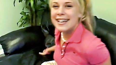 Young petite blonde smiling happily in tight pink polo shirt on leather couch