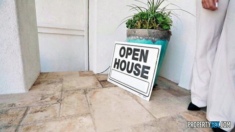 Open house sign leaning by white modern home entrance with potted plant and realtor's white pants visible