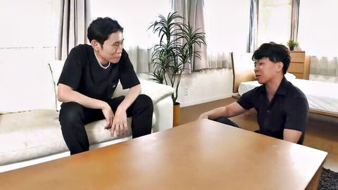 Two clothed Asian men sitting and chatting across table in living room