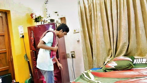 Young South Asian man in white shirt standing by red fridge in bedroom with bed and curtains