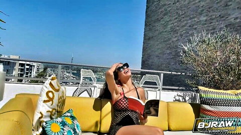 Tanned brunette MILF with big tits in skimpy bikini reads book on sunny rooftop
