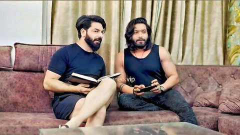 Two bearded Indian men chilling on couch, one reading book other holding phone controller