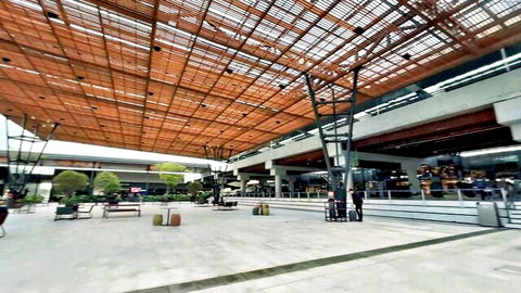 Spacious modern airport plaza with wooden slat roof, white floors, benches, plants and distant people.