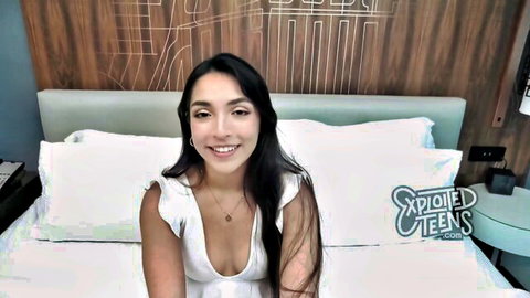 Petite brunette teen smiling with deep cleavage in low-cut white top on hotel bed