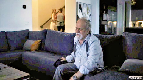 Older bearded man in suspenders sits on couch holding remote in living room with couple on stairs