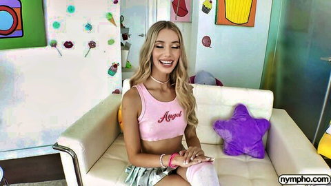 Blonde babe in pink Angel top and skirt, smiling playfully on couch