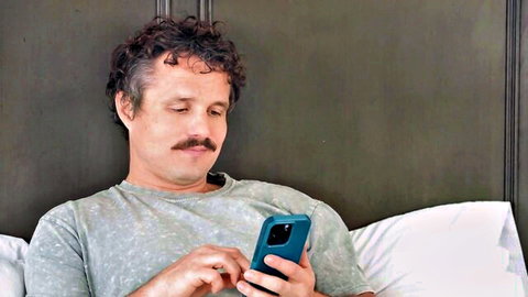 Curly-haired man with mustache checks phone in bed wearing gray t-shirt