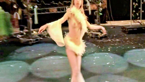 Topless blonde babe with perky tits poses arms outstretched in feathery yellow costume on stage