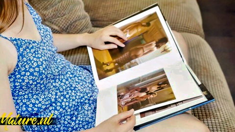 Young redhead in blue floral dress sits on couch browsing open photo album of busty nude women flashing tits and pussies