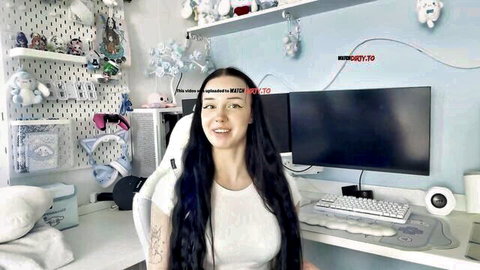 Smiling tattooed teen gamer girl in white top at streaming desk with plush toys