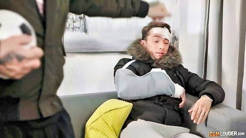 Tattooed injured man with head bandage and arm sling relaxes on couch while buddy in green jacket stands behind holding ball