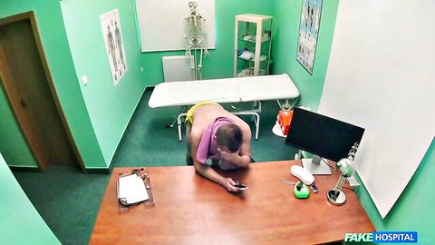 Bald man in yellow shorts and towel sits on hospital floor at desk checking phone in medical office