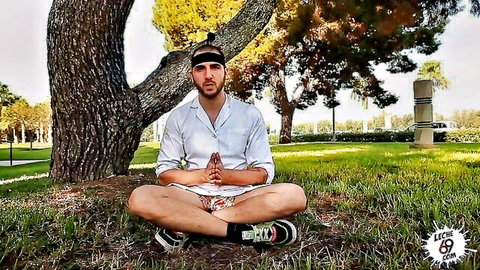 Attractive man meditating zen style in shorts and sneakers on grass under tree