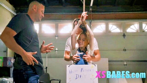 Petite redhead teen smiles holding Happy Father's Day sign with muscular DILF stepdad in naughty taboo garage tease