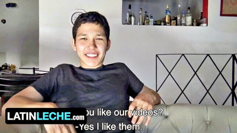 Young Latino guy smiling in gray t-shirt on couch teasing for LatinLeche gay porn