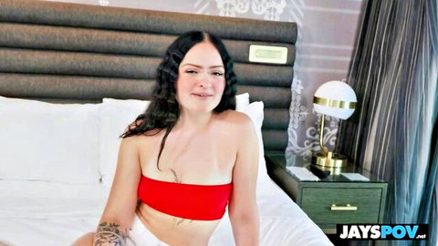 Skinny young brunette with small tits smiles on hotel bed in tight red tube top and white shorts POV