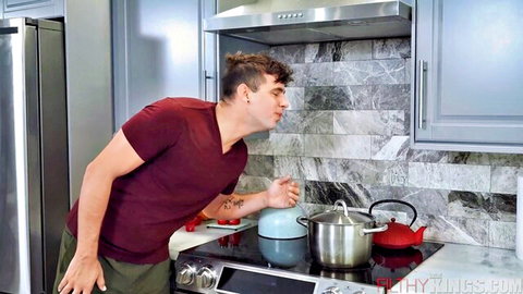 Handsome young man leaning over stove checking pots and kettles in modern gray kitchen