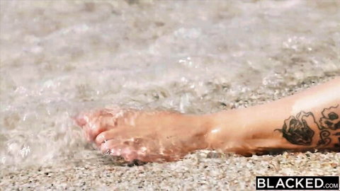 Rae Lil Black's tattooed foot in shallow beach water, teasing view
