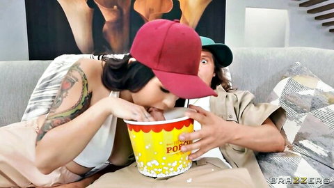 Tattooed brunette babe in red cap playfully buries face in popcorn bucket held by guy