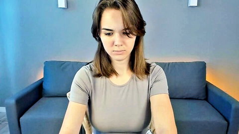 Brunette webcam girl pouting seductively on blue couch in tight gray t-shirt