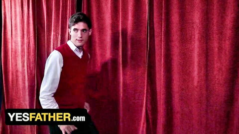 Handsome young stud in red vest and white shirt poses seductively before red curtains in gay taboo priest roleplay