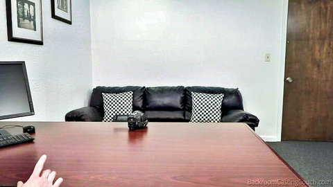 Empty backroom casting couch room with black leather sofa, desk, computer, and camera ready for amateur auditions