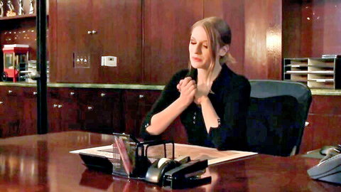 Ela Darling in black top, hands clasped, intense gaze in office scene