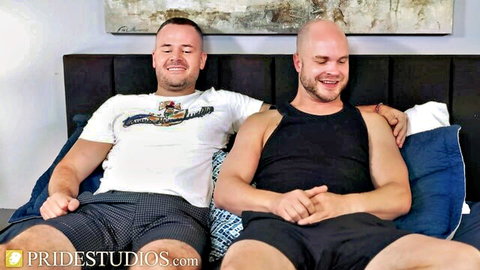 Athletic brunette and bald gay studs smiling on bed in shorts and tanks