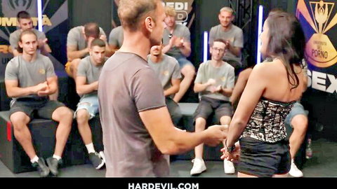 Sexy woman in black corset and mini skirt surrounded by clapping muscular men in Hardevil studio show