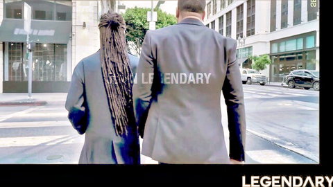Ebony woman with long dreadlocks in tight dress walks hand-in-hand with suited man on urban street