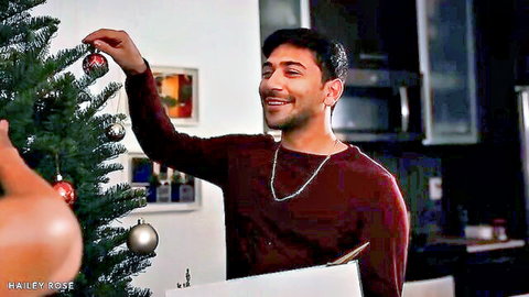 Smiling young guy in maroon shirt hangs ornament on Xmas tree with babe's curvy tan ass teasing foreground