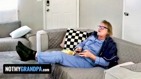Mature blonde grandma in blue scrubs lounging on couch eating snacks with game controller